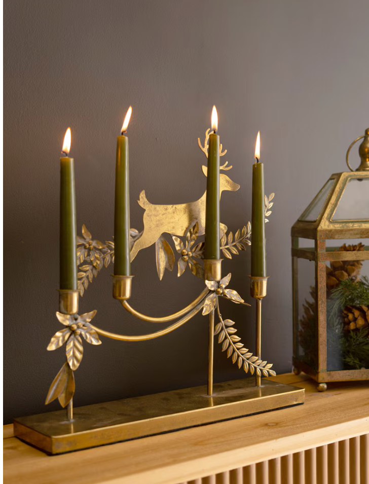 A brass candelabra with a deer silhouette and leafy branches, holding four green taper candles, displayed on a wooden shelf against a dark gray wall.