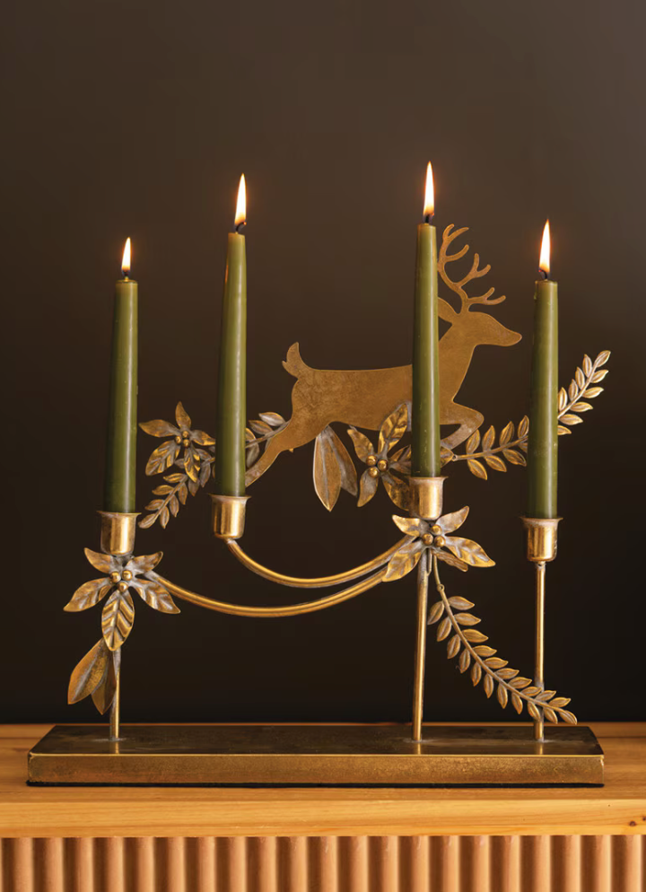 A decorative metal candelabra with four lit green candles, featuring a stag silhouette and leafy scrollwork in a warm gold/brass tone, displayed against a dark background.