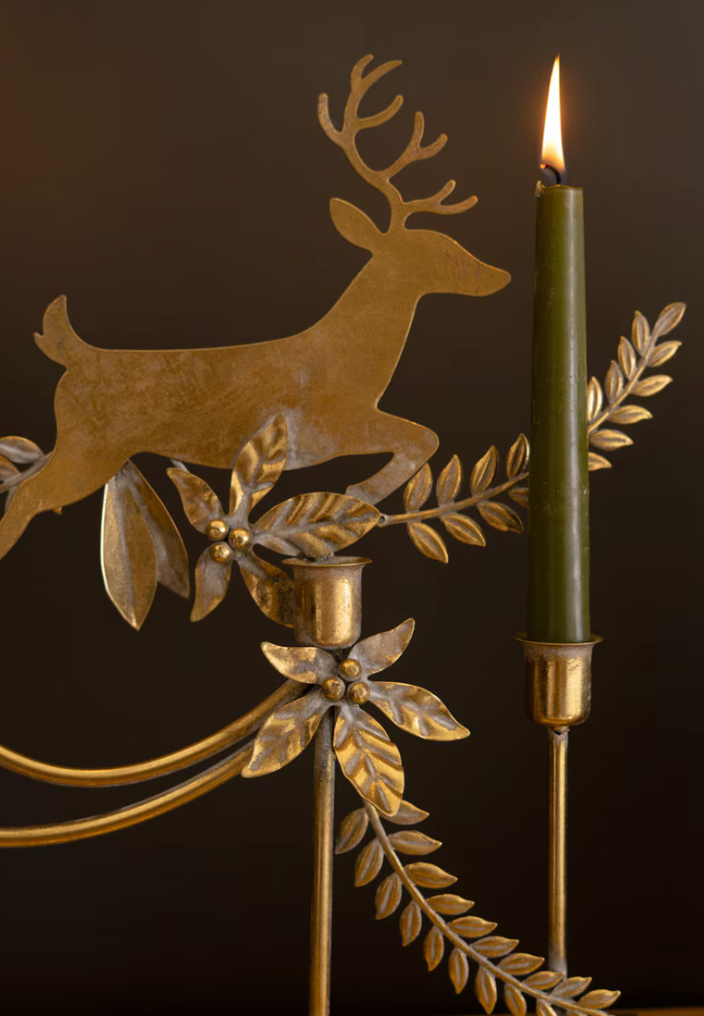 A close-up of a decorative brass holiday candelabra featuring a running deer silhouette, leafy branches, and a lit dark green taper candle against a dark brown background.