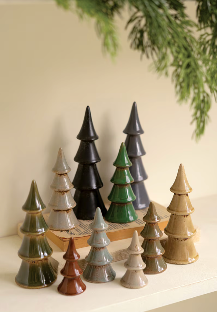A set of ceramic Christmas tree ornaments in various colors including green, black, brown, beige, and gray, arranged on a light-colored surface with a book and pine branches in the background.