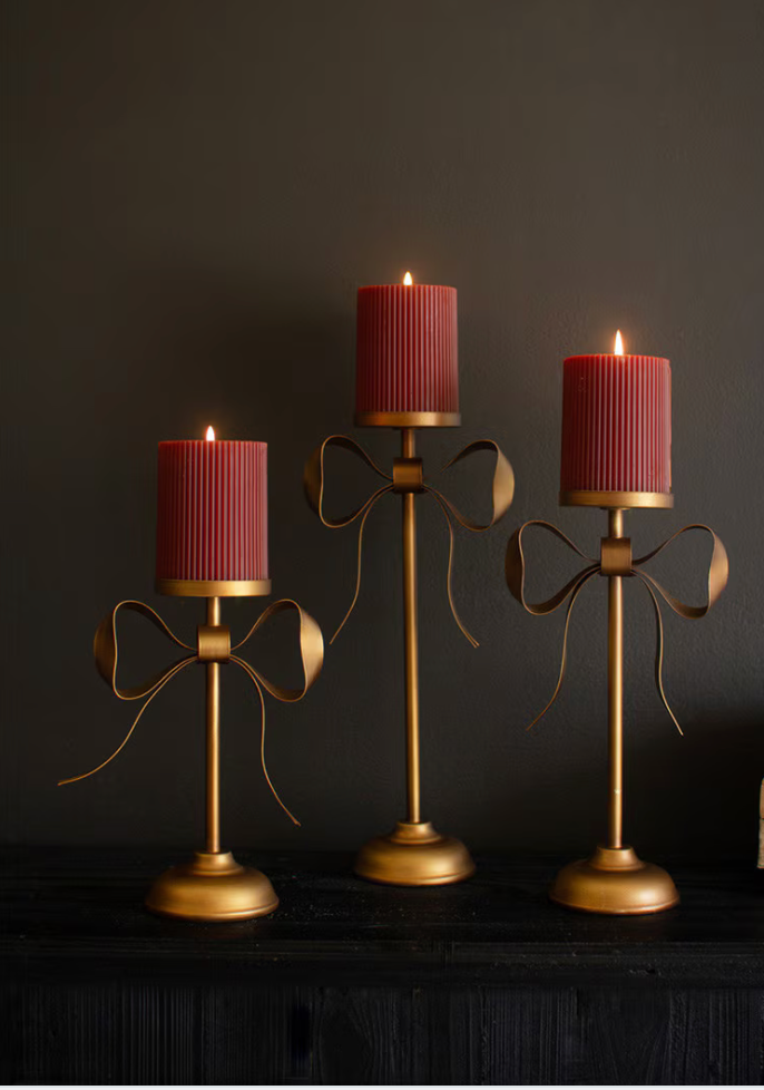 Three gold metal candle holders with ribbon-like curved stems and cylindrical ribbed red candles, displayed against a dark background.