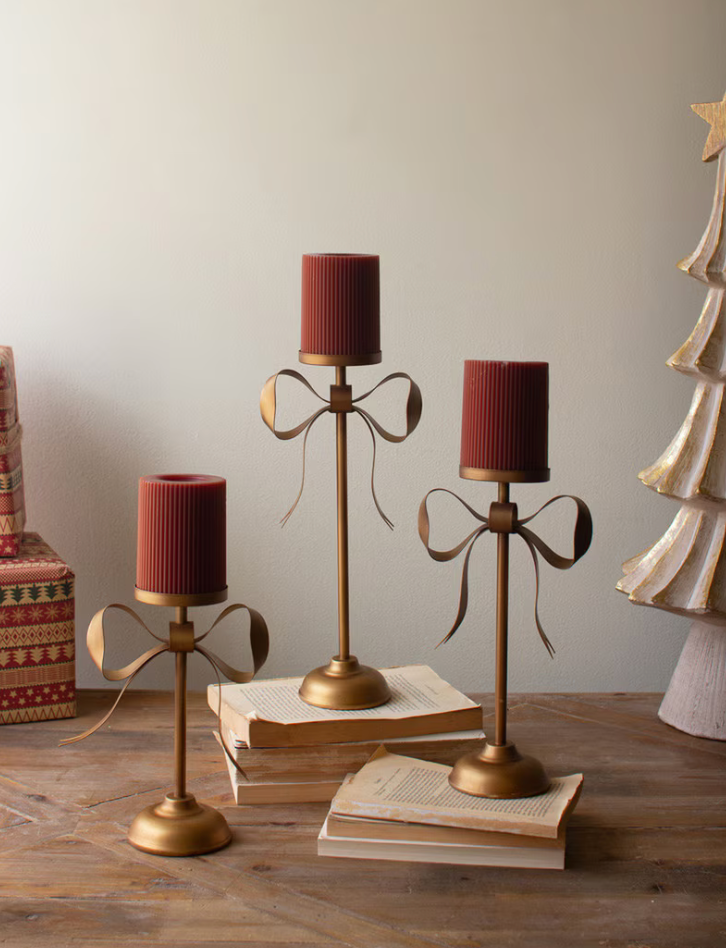 Three gold-toned bow-shaped candlesticks with ribbed dark red cylindrical candle holders, styled on a wooden table with vintage books and a Christmas tree decoration against a neutral wall.