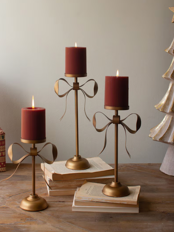 Three gold-toned metal candlestick holders with ribbon-like loops are arranged on stacked books on a wooden table. Each holder supports a ribbed dark red pillar candle.