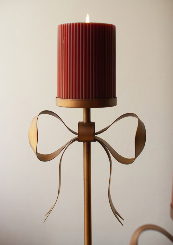 A ribbed red pillar candle in a flared brass holder with a decorative bow-shaped metal accent, shown lit against a plain light background.