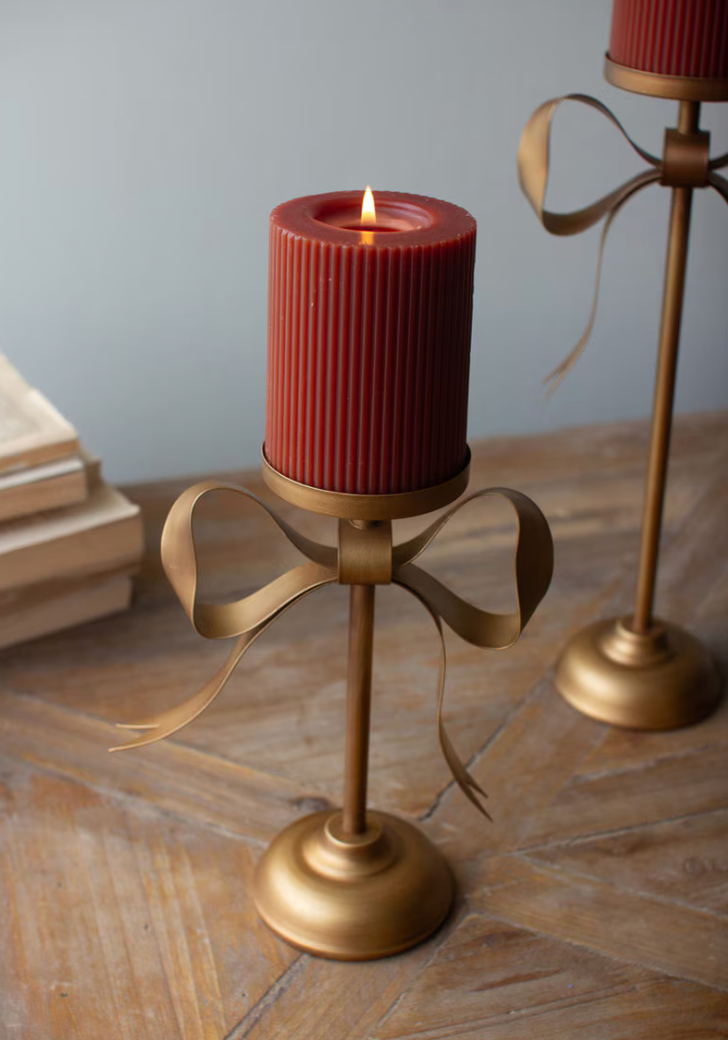A gold bow-shaped metal candle holder with a lit red ribbed pillar candle, set on a wooden table with a blurred second matching holder in the background.