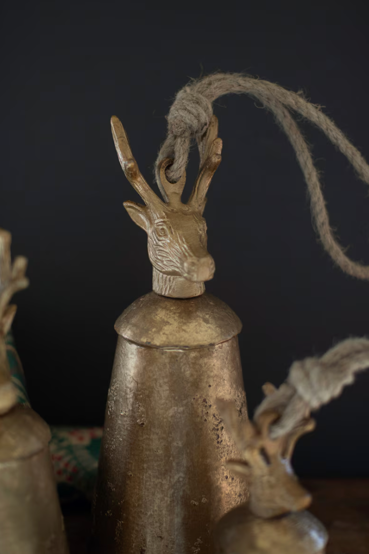 A rustic brass bell with a carved deer head finial and a knotted rope handle, shown against a dark background with other similar bells partially visible.