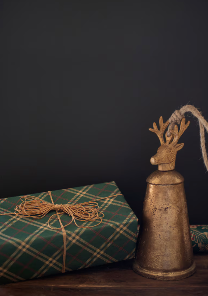 A rustic holiday still life with a wrapped gift box in green plaid paper and twine beside a vintage brass bell topped with a deer head, set against a dark charcoal background.