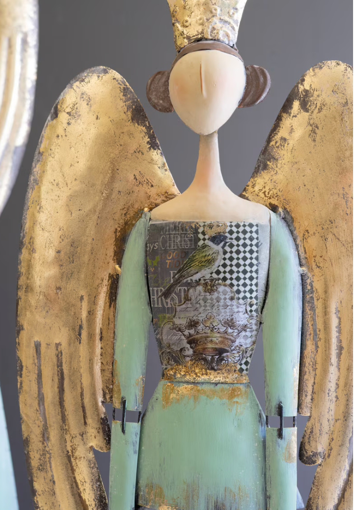A stylized angel figurine with large distressed gold wings, a pale face and neck, a mint green dress, and a patterned collage-style bodice featuring a bird and checkerboard print, shown against a dark gray background.