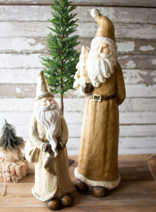 Two rustic Santa Claus figurines, one large and one small, standing on a wooden surface in front of a distressed white wood wall with a small green tree behind them.