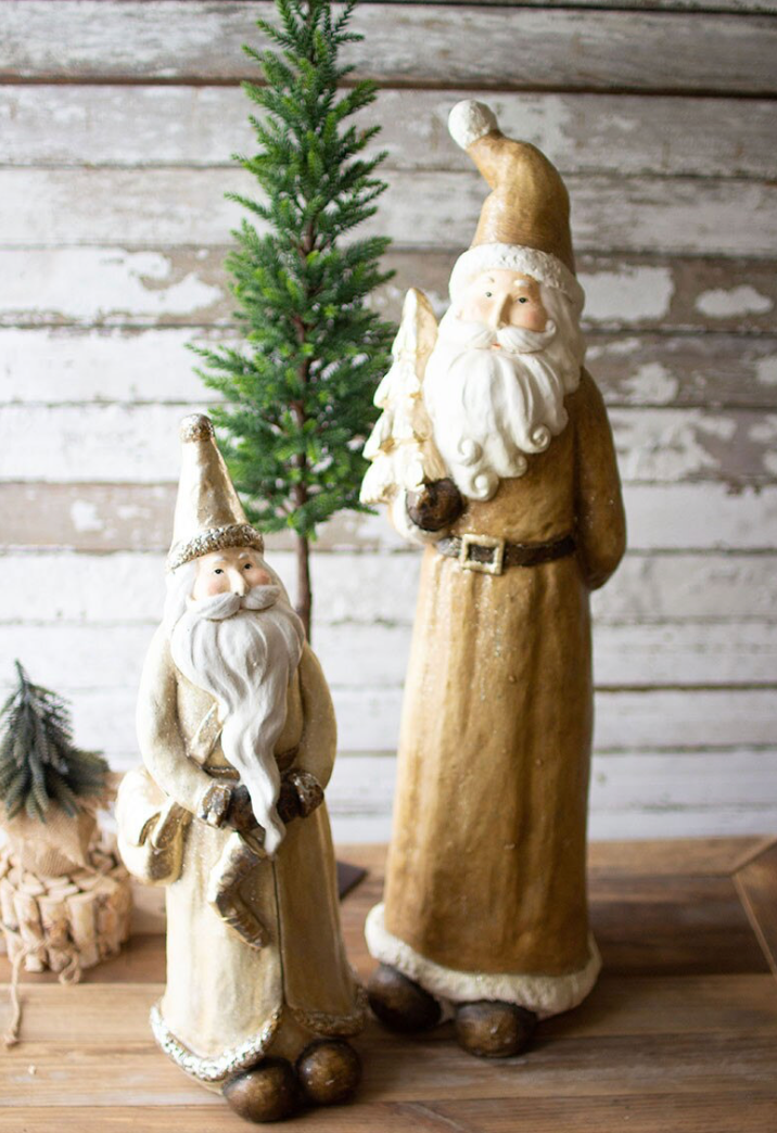 Two rustic Santa Claus figurines, one large and one small, standing on a wooden surface in front of a distressed white wood wall with a small green tree behind them.