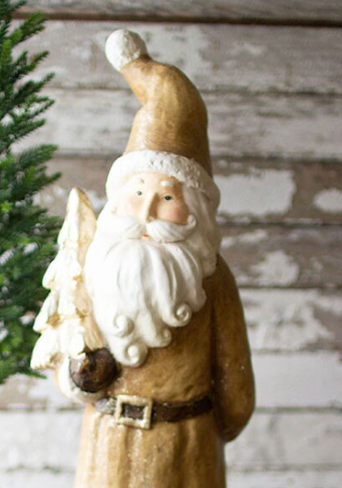 A rustic Santa Claus figurine with a tall tan hat, white beard, and brown robe, holding a small white tree, set against a distressed white wood background with a green pine branch on the left.