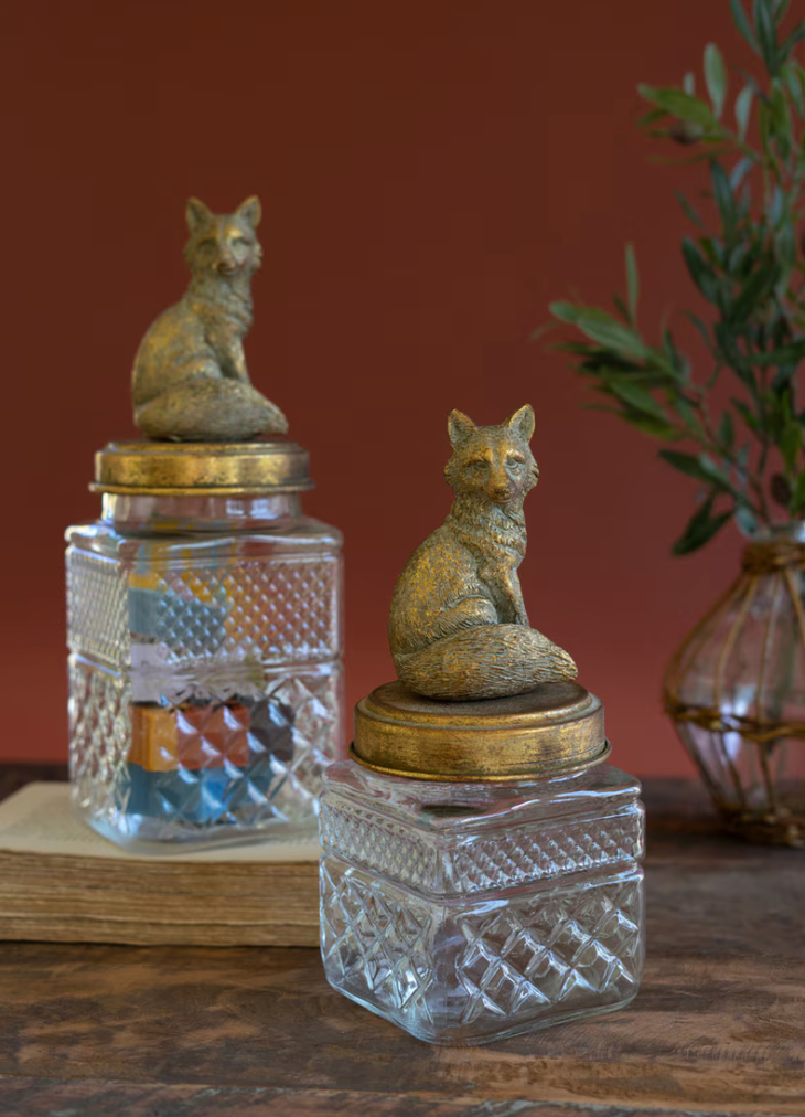 Two vintage-style clear glass jars with textured diamond-cut sides and gold-toned lids, each topped with a small seated fox figurine, displayed on a wooden surface against a warm reddish-brown background.