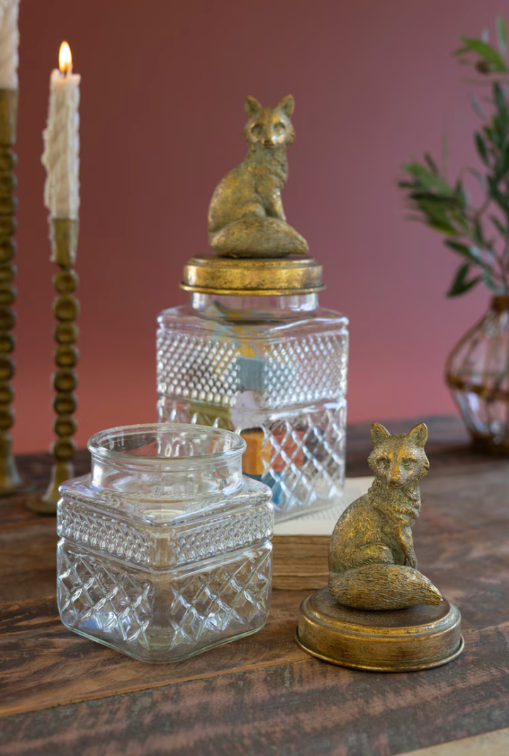 Two decorative glass jars with textured diamond-cut patterns, one topped with a bronze fox figurine and the other holding a lit candle, displayed on a wooden table against a dusty rose background.