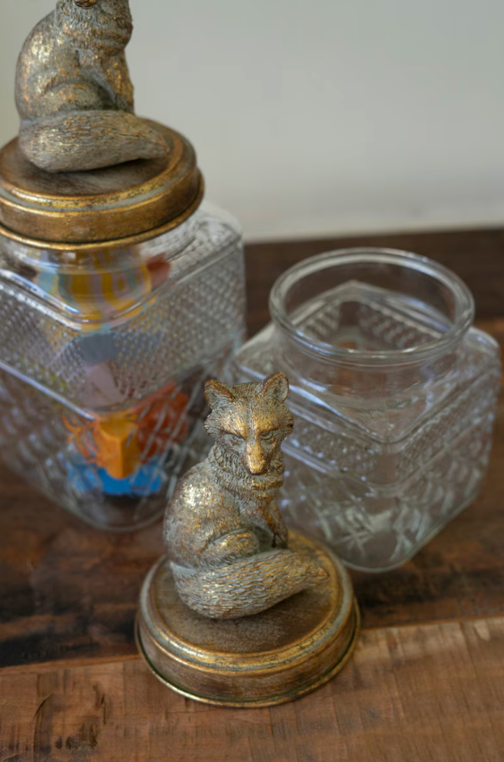 Decorative metallic fox figurines displayed on a wooden surface, including one on a round base in the foreground and another atop a glass jar lid in the background.