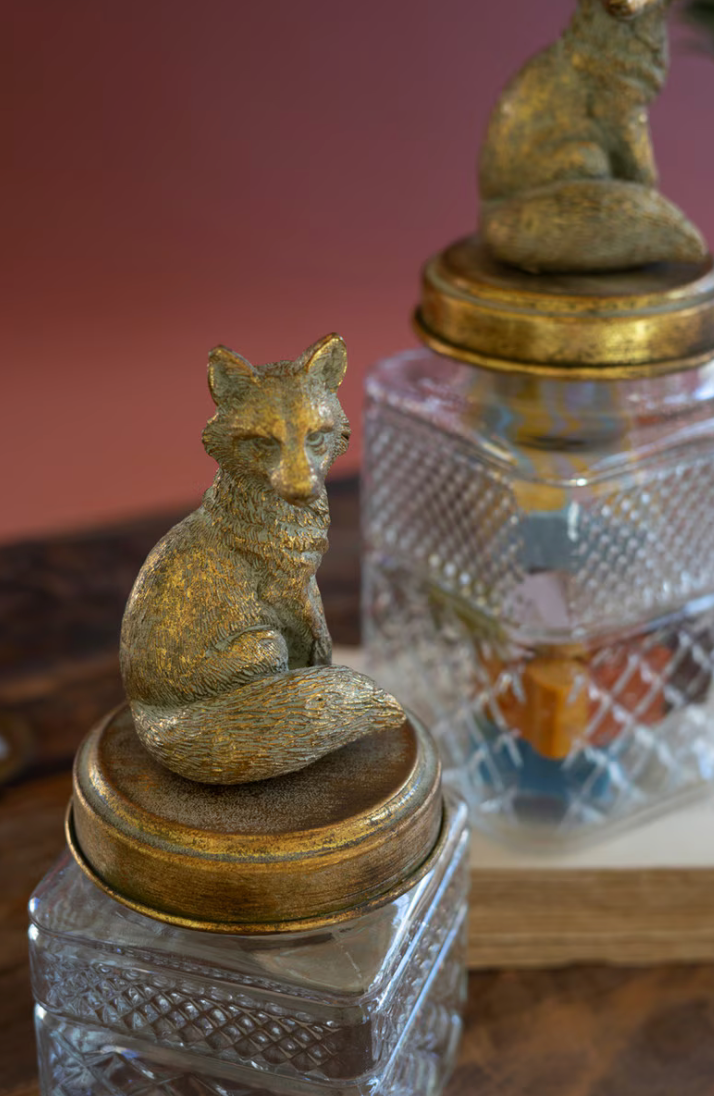 A close-up of a decorative gold-toned fox figurine perched on top of a clear textured glass jar with a round gold lid, with a second matching jar blurred in the background against a warm reddish-brown backdrop.