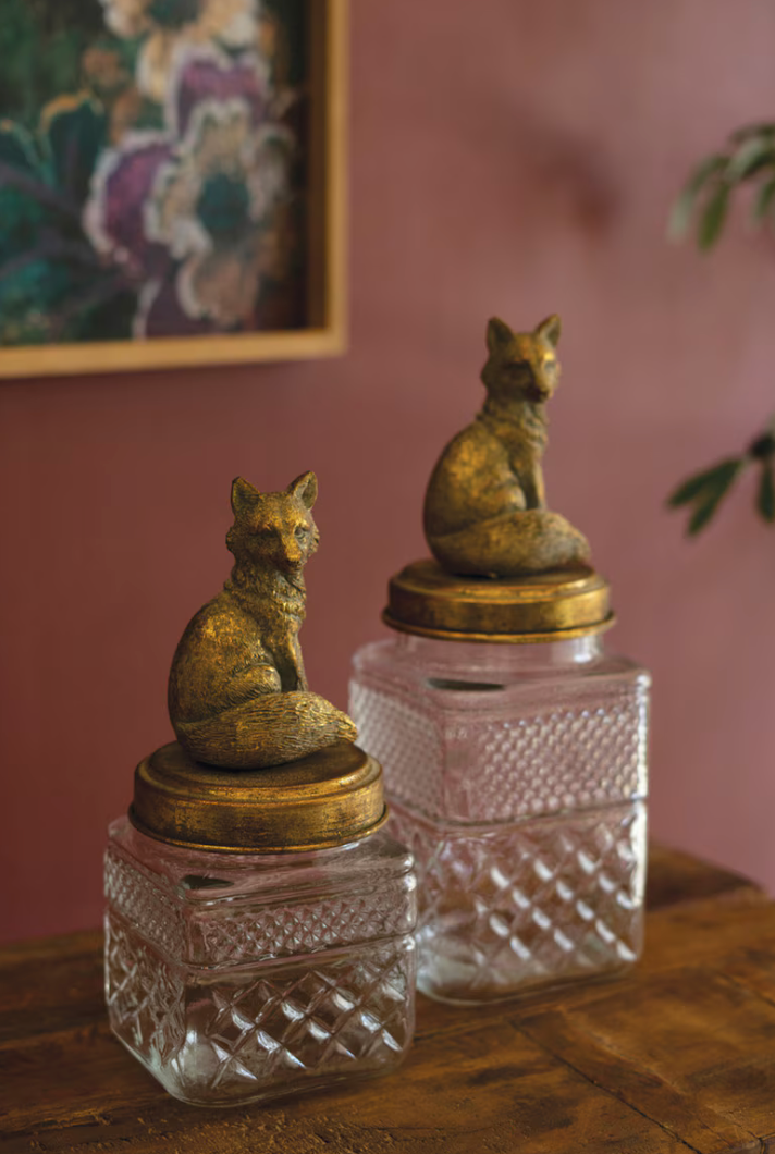 Two vintage-style glass money boxes with gold fox figurines on top, displayed on a wooden table against a dusty rose wall.
