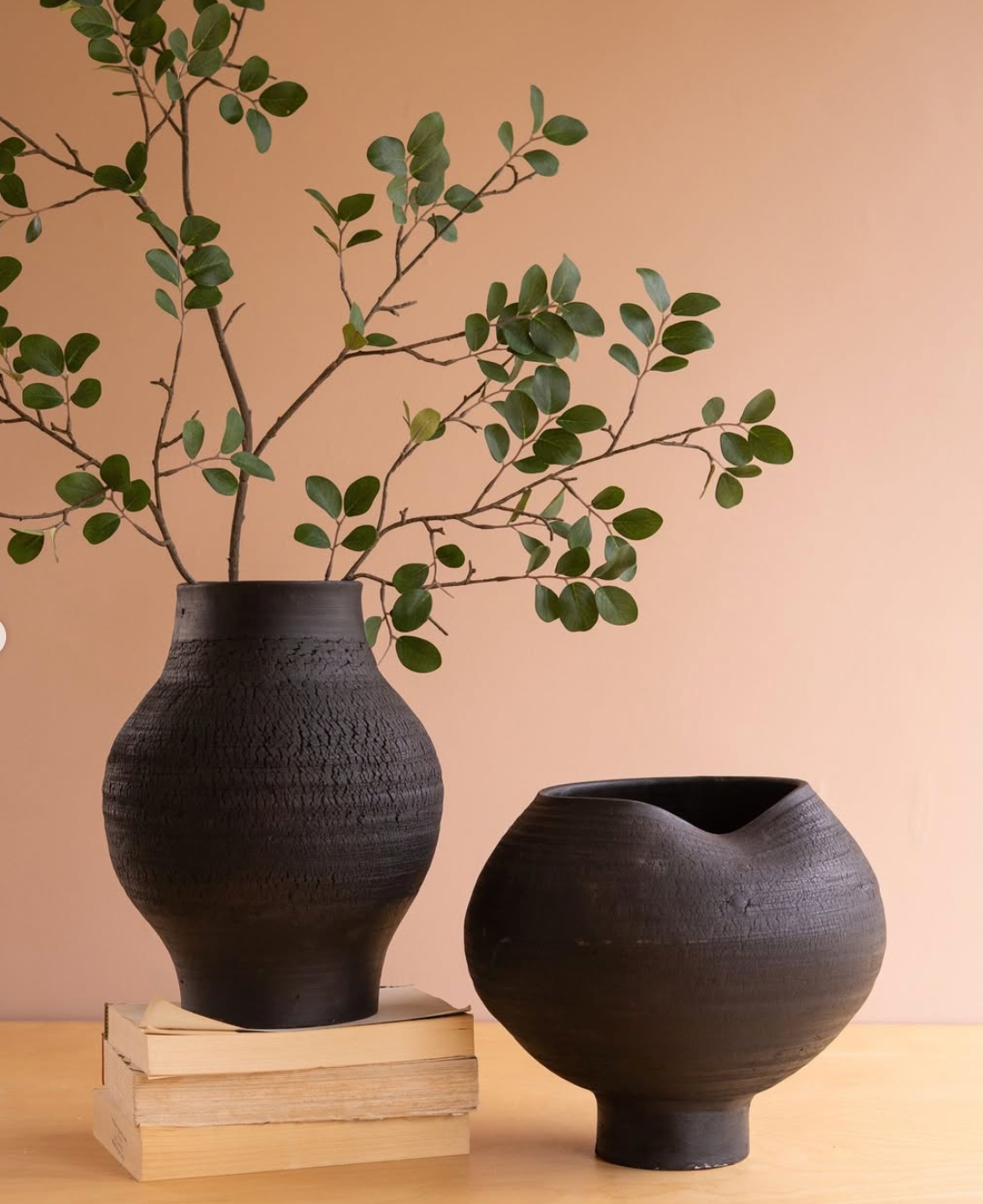 Two matte black ceramic vases on a light wood table against a warm peach background. The taller vase on stacked books holds leafy green branches, and the shorter vase sits beside it.
