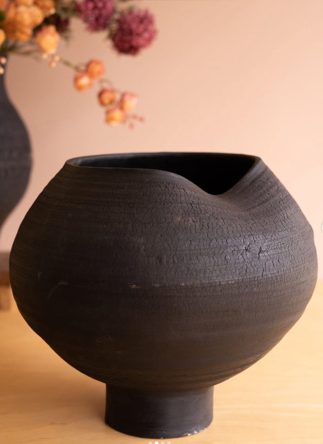 A matte black ceramic vase with a textured, crackled surface and a wide, rounded body on a short pedestal base, set on a light wooden surface against a warm beige background.