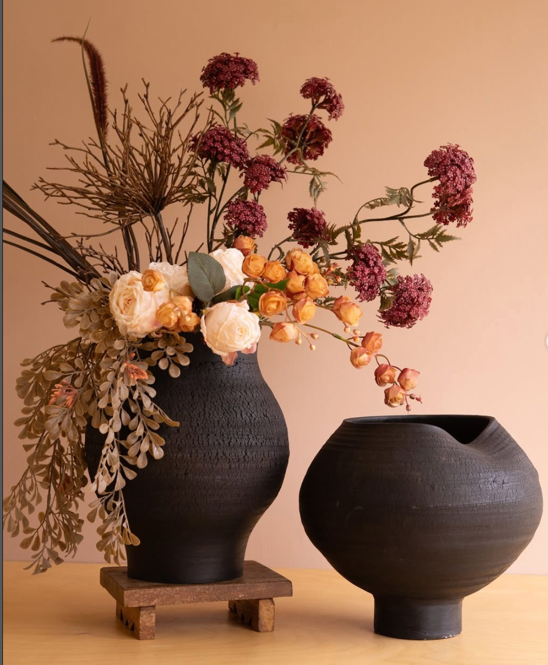 Two matte black ceramic vases displayed on a light wood surface against a warm beige wall. The taller vase sits on a small wooden stand and is filled with dried botanicals, burgundy flowers, and cream roses.