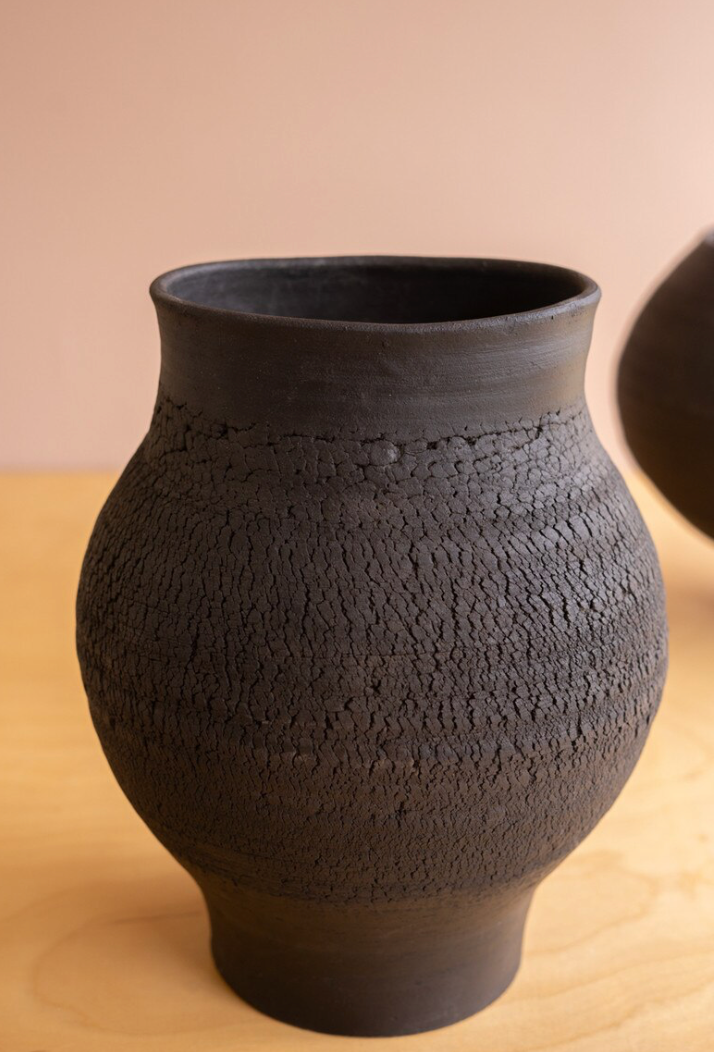 A dark charcoal-colored ceramic vase with a textured, crackled surface and a flared rim, shown on a light wood surface against a warm beige background.