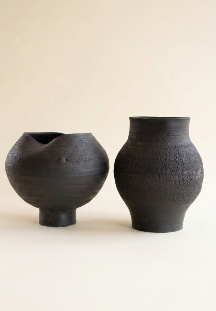 Two handmade dark charcoal ceramic vases with textured surfaces, one with a small spout and one with a flared base, set against a solid warm beige background.