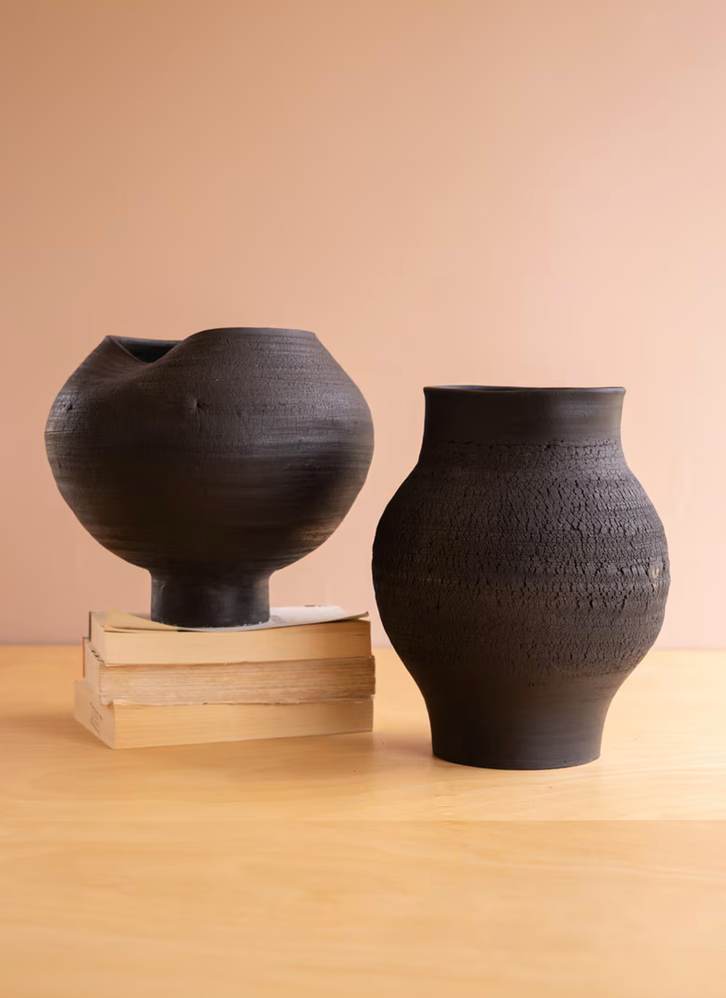Two dark textured ceramic vases displayed on a light wood table against a warm beige background. One vase is black with a wide flared rim and sits on stacked books, while the other is a dark brown textured vase standing next to it.