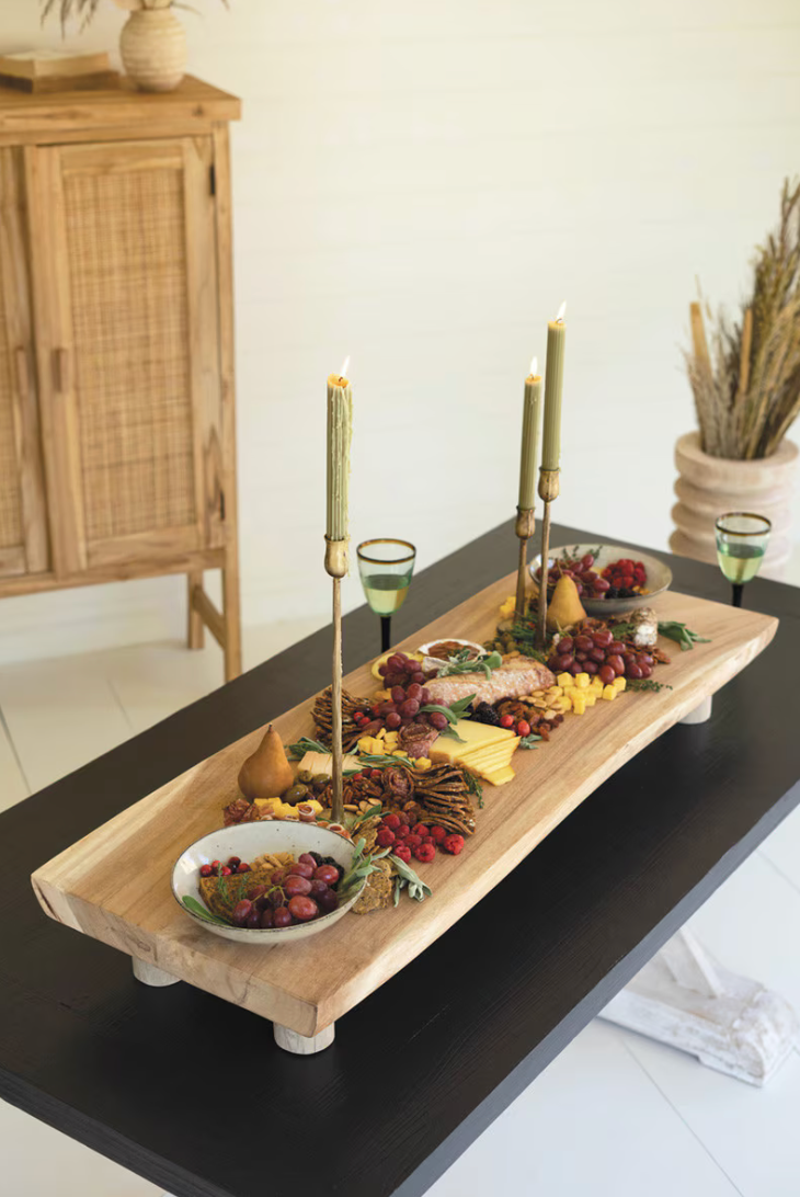 A long wooden serving board styled with food, three tall candlesticks, and small bowls on a black table in a bright indoor setting.
