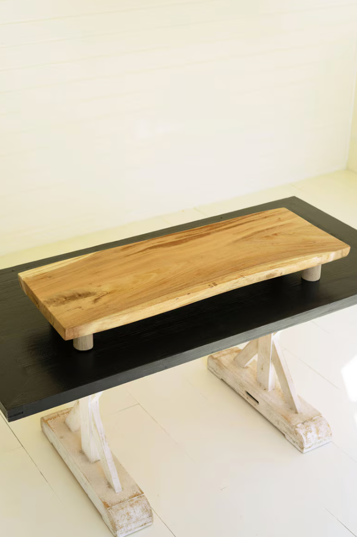 A rustic live-edge wooden table top with a natural light brown grain, mounted on a black rectangular board supported by distressed white trestle-style legs, set against a cream paneled wall.