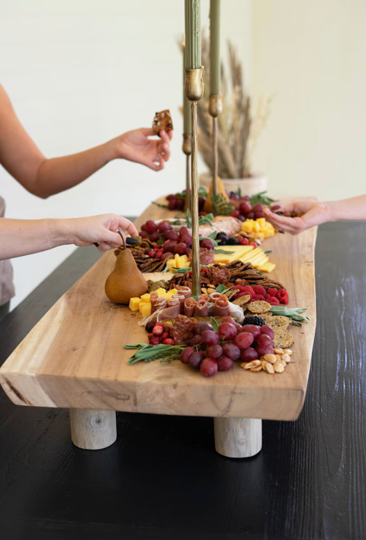 A large rectangular wooden charcuterie board with a natural live-edge style sits on a dark table, surrounded by food items like grapes, pears, nuts, and cheeses. Multiple hands are visible reaching in from both sides, and a decorative dried plant arrangement is in the background.