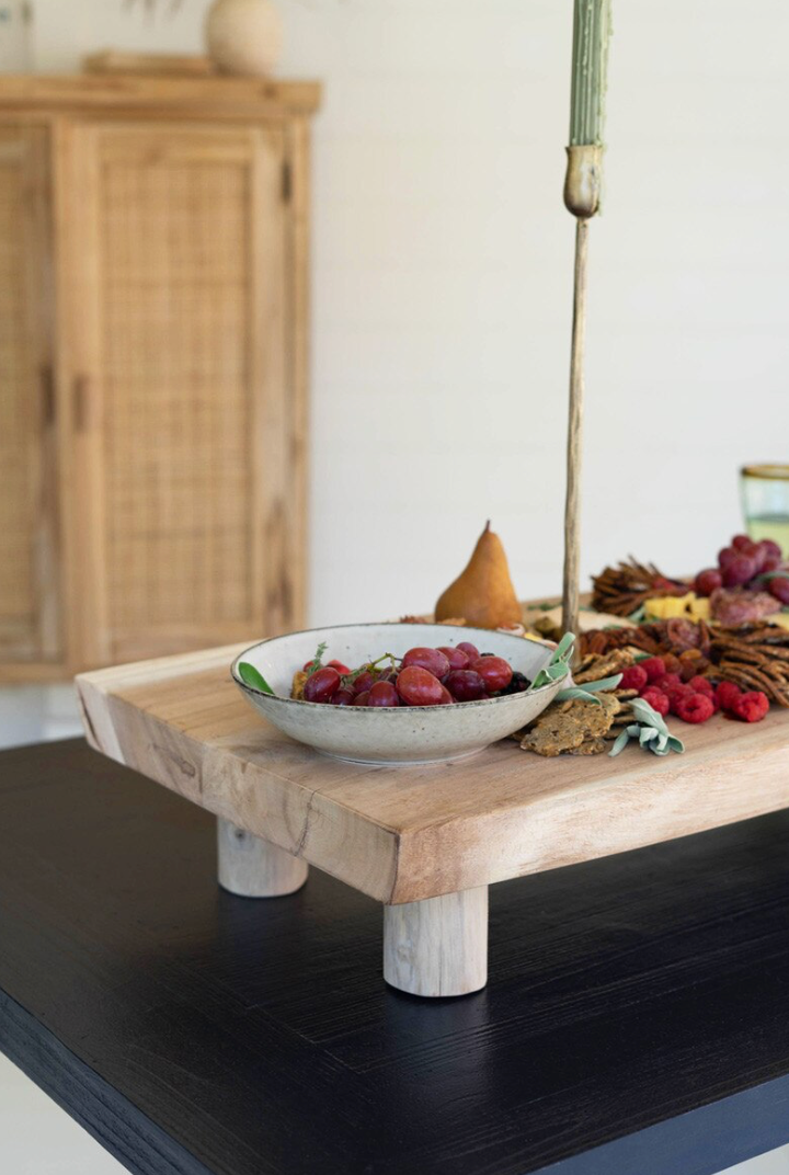 A rustic wooden serving board with chunky legs, styled with a bowl of red grapes, a pear, and dried fruit on a black table in a bright room.