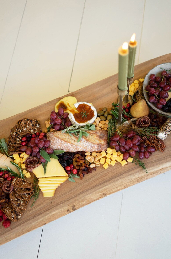 A rustic holiday charcuterie board arranged on a long wooden serving board, featuring sliced cheese, a stuffed bread roll, grapes, nuts, dried fruit, candles, and small decorative rose arrangements on a light tabletop.