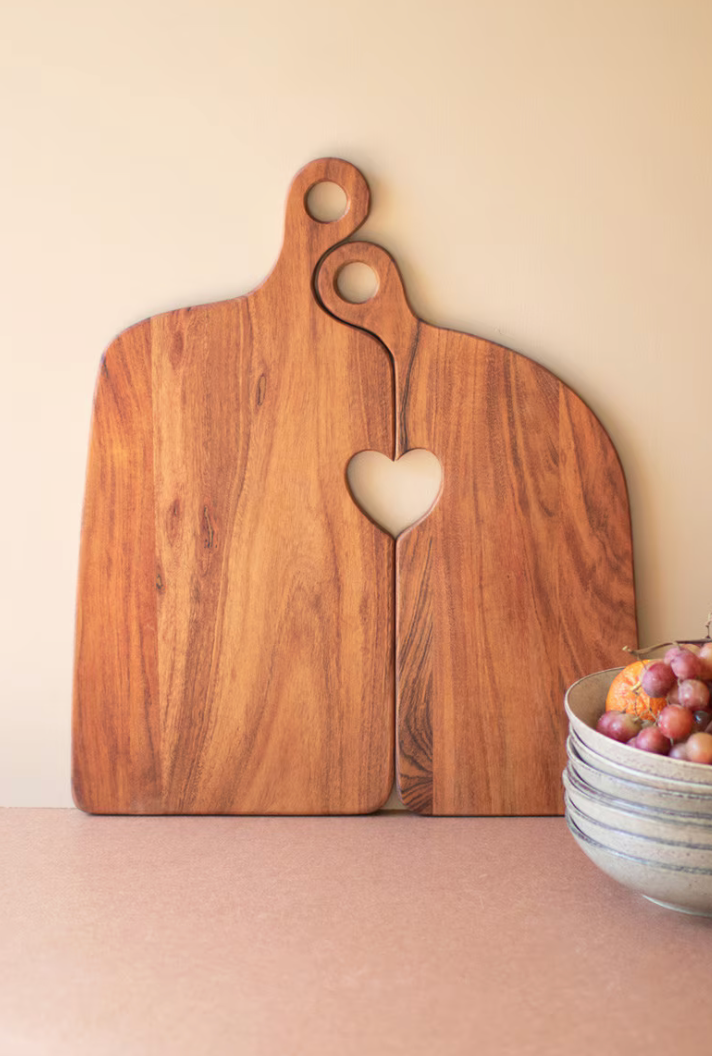 Two wooden serving boards with a heart-shaped cutout in the center, leaning against a beige wall on a pink surface, with a small stack of bowls and fruit at the right edge.