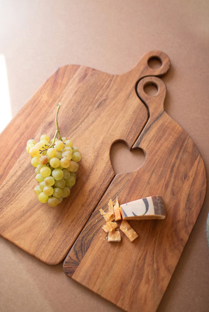 Two wooden serving boards with a heart-shaped cutout and loop handles, styled with a bunch of green grapes and a wedge of marbled cheese on a warm beige surface.