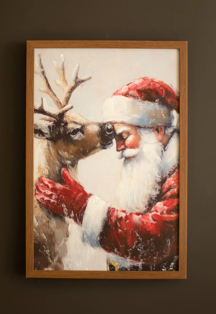  "Framed Santa with Deer Painting by Kalalou showing Santa in red coat and gloves tenderly embracing a woodland reindeer in a layered paint-over-print style, in warm oak frame against dark wall"