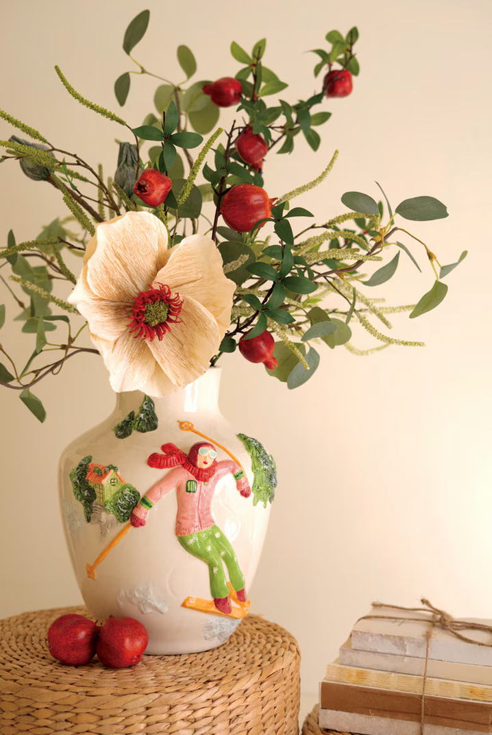 Ceramic Christmas Vase with Skier by Kalalou filled with red berry stems, eucalyptus, and cream poppy bloom on woven rattan stool with red apples, cream background"