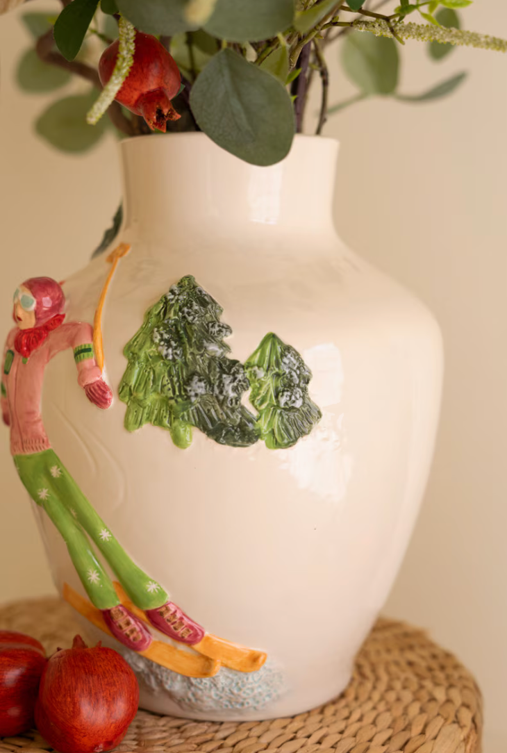 "Side detail of Ceramic Christmas Vase with Skier showing hand-sculpted skier in pink jacket and green pants with snow-dusted pine trees in raised relief on white ceramic"