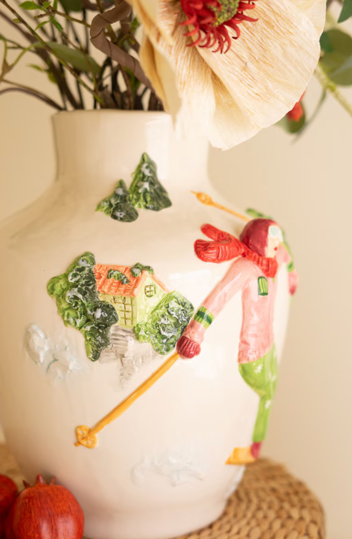 "Close-up of Ceramic Christmas Vase with Skier showing raised relief skier with poles, alpine cottage, and snow-covered pine trees on glossy white ceramic with holiday florals above"