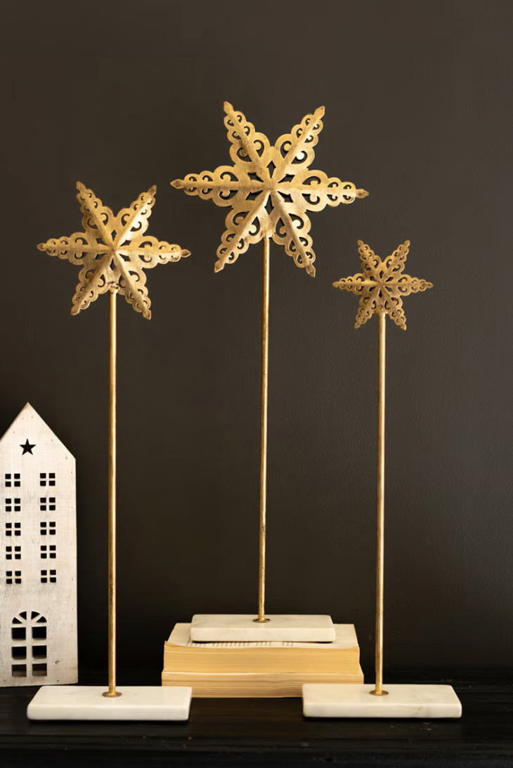 set of 3 metal gold stars with long metal sticks in a marble stand with a dark background and a small white clay house decor to the left. 
