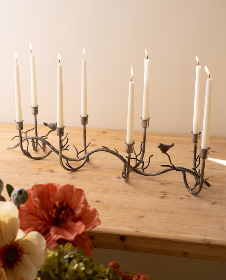 A rustic metal candelabra with nine branches, small bird details, and lit white taper candles, displayed on a light wood table with flowers in the foreground.