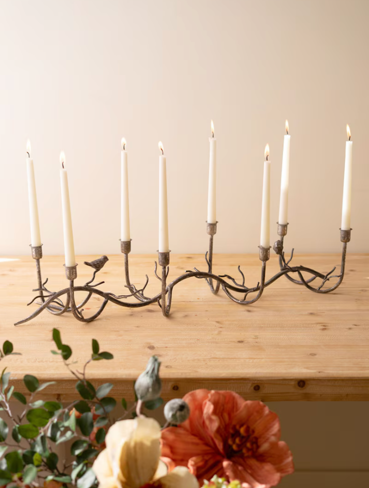 A rustic nine-light candle holder made from dark branches, decorated with small bird figures and holding nine white taper candles, displayed on a light wood shelf against a beige wall.