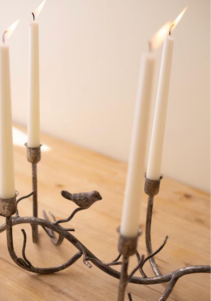 A rustic metal candelabra with a bird perched on the branches, holding several lit white taper candles on a light wood surface against a soft beige background.