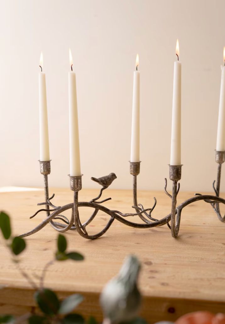 A rustic metal candelabra with five candle holders shaped like branches and a small bird figure in the center, holding white taper candles on a light wood surface against a soft beige background.