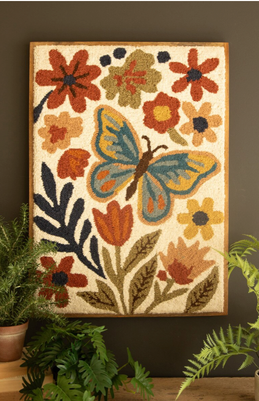 A framed hand-hooked tapestry featuring a large blue and yellow butterfly surrounded by orange, red, and green flowers on a cream background, hung on a dark brown wall with potted plants below.