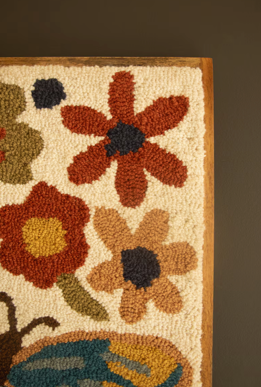 A close-up of a hand-tufted or punch needle rug showing a floral pattern with textured loops of orange, red, tan, blue, olive green, and cream on a dark brown background.
