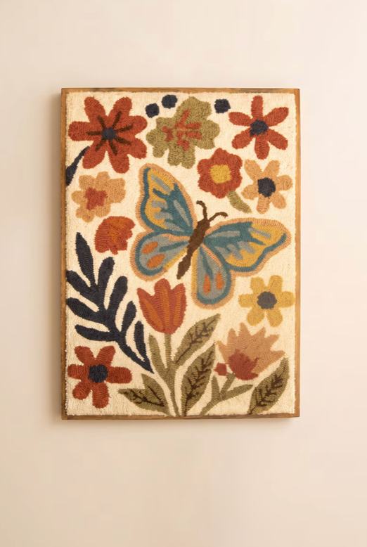 A framed hand-tufted wall hanging featuring a large blue and yellow butterfly surrounded by red, orange, and yellow flowers on a cream background, mounted on a light beige wall.