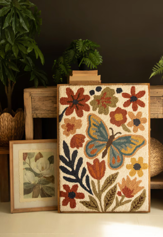 A rectangular woven rug with a floral and butterfly pattern, displayed on a rustic wooden bench in a styled room setting with plants and a framed print.