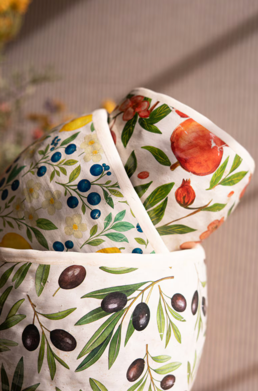 Three fabric-covered canisters or baskets with a white base and colorful botanical prints, including olive branches, blue berries, yellow flowers, and a red pomegranate, arranged together on a neutral background.
