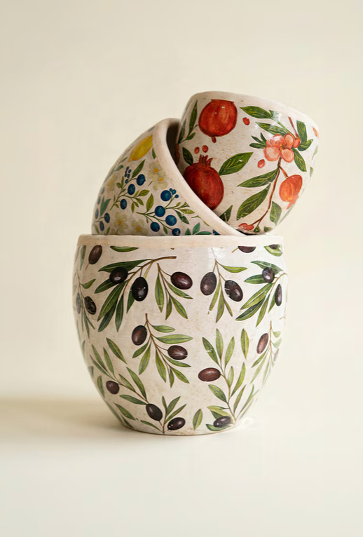 Three stacked ceramic bowls with hand-painted botanical and fruit motifs, including an olive branch pattern on the front bowl and pomegranate and berry designs on the stacked bowls behind it, set against a warm cream background.