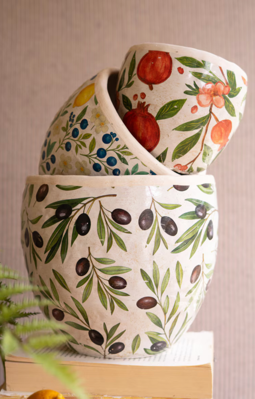 Three stacked ceramic planters with a textured cream glaze, decorated with hand-painted botanical motifs including green olive branches, red pomegranates, blue berries, and orange fruits, displayed on a book with a blurred plant in the foreground.