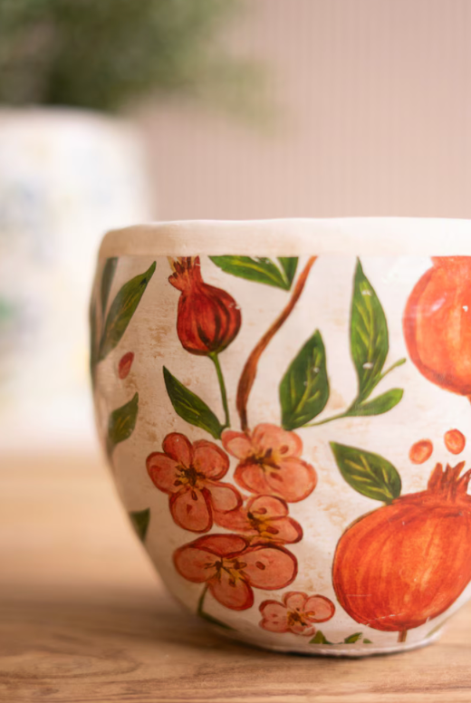 A close-up of a ceramic planter pot decorated with hand-painted orange pomegranates, green leaves, and peach blossoms on a cream-colored background, set on a wooden surface.